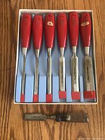 Six red-handled chisels aligned in a box with metal blades visible, plus one separate single chisel with wooden handle placed below.