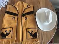 Cowboy vest in mustard yellow with black moose patches, horseshoe designs, and fringe, lying next to a white cowboy hat.