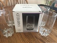 Four large crystal drinking glasses standing on a wooden table, box displayed behind them showing the brand Fairfield and product image.