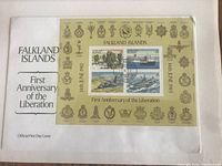 Photo of Falkland Islands official first day cover commemorating the First Anniversary of the Liberation showing four postage stamps with naval and military scenes surrounded by gold-toned military insignias on the cover.