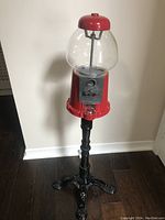 Full view of the red metal and glass gumball machine mounted on the black metal stand showing the entire unit from top to feet.