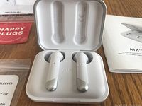 Happy Plugs Air 1 wireless earbuds in open white charging case, displaying earbuds with metallic silver accents, on wooden surface.