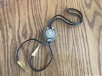 Full view of the bolo tie showing braided black cords with gold tips and oval medallion with bull head and horseshoe design.