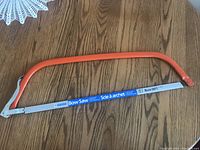 Metal bow saw with orange painted frame and attached blade laid on wooden floor.