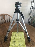 Full view of the tripod standing on a wooden table with the instruction sheet placed underneath it. Shows aluminum legs and black mount head.