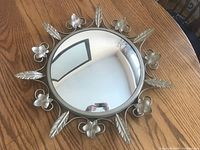 Top view of the vintage convex round wall mirror on a wooden table, showing the decorative fleur-de-lis and leaf metal frame with silver finish.