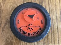 Close-up photo of the Pow-R-Puck showing an orange dial with numbers 0-100 and resistance adjustment between Low and High, surrounded by a black rubber tire-like rim.