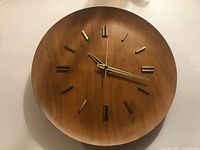 Front view of the round teak wood wall clock showing hour markers, hands, and surface with minor chips near number 5.