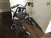 Front angled view of the red metal frame walker with white wheels and black seat with crack visible, folded handle grips with hand brakes.