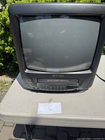 Front view of a compact black CRT TV with integrated VHS player below the screen, showing controls and screen.