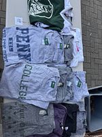 Top view of folded T-shirts showing multiple gray 'PENN COLLEGE' printed shirts, plus other light colored tees in a pile outdoors.