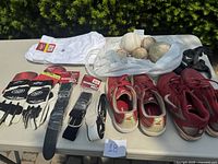 Wide view of the lot showing baseball pants, gloves, belts, baseballs in bag, and cleats on table outside.