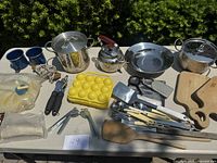 Wide view of all camping kitchen items laid on table, showing pots, pans, utensils, egg tray, mugs, kettle and cutting boards