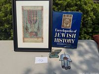 Photo of the collection showing the framed menorah artwork, Encyclopedia of Jewish History book, metal menorah wall decoration, and stained glass Hamsa hand hanging.