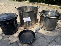 Assorted used large pots including black enamel soup pot, large stainless steel pot, stainless steel pot with strainer insert, and black frying pan.