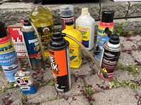 Photo shows assorted cleaning sprays, oils, and antifreeze containers placed on outdoor stone surface. Includes various brands and types of motor and household fluids.