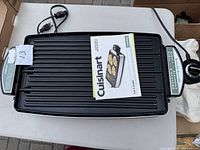 Full top-down view of the Cuisinart electric grill showing the reversible cooking surface and attached temperature control dial with cord alongside instruction manual placed on grill.
