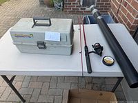 The tackle box closed beside the fishing rod, rod case, and spool of line on a white folding table outdoors.