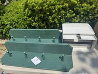 Two green wooden hook racks with rounded pegs and one white aluminium paper towel storage box against greenery background.