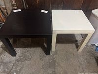 Image showing two square Ikea Lack end tables side by side, one black and one white, placed on concrete floor.