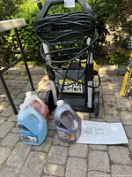 Yellow and black Karcher power washer with power cord wrapped around the handle, accompanied by three gallon-sized bottles of wood cleaning compounds in blue, red, and dark colors on stone pavement.