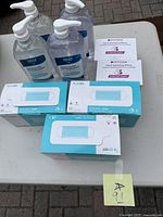 Four large pump bottles of clear hand sanitizer liquid, two boxed packages of ASTM Level 2 blue procedure masks, and two packets of Hygia hand sanitizing wipes on a white table outdoors.
