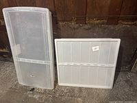 Two large plastic storage boxes shown standing upright, one with lid removed and leaning against wall