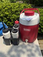 Thermos jug with spigot and five drinking bottles in black plastic caddy with handle outdoors