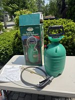 Photo showing the lawn and garden sprayer, original box, and instruction sheet on a table outdoors with greenery in the background.
