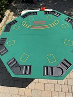 Top-down image showing green felt poker table top with yellow card outlines and red/yellow centered text. Black molded plastic trays at each player position with slots for chips and cards plus cup holders.
