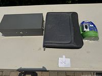 Safety deposit box, leather notebook, and iGo charger shown together on a beige surface with a paper numbered 24.