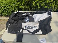Front and side views of a black and white Toronto Blue Jays sports bag displaying team logos, placed on a table outside with greenery in the background.
