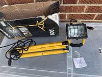 Yellow LED tripod stand workshop light with plug and cord laid out with its original box in the background. The tripod has foldable legs and a protective wire grill on the lamp.