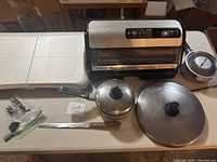 Wide shot showing FoodSaver vacuum sealer, white cutting board, pots with lids, and knife arranged on white surface.