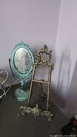 Oval antique mirror with turquoise ornate metal frame and ornate metal picture stand on table against wall.