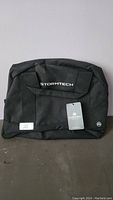 Front view of black Stormtech tech bag featuring brand logo and attached tag, placed against a wall.