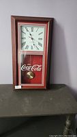 Front view of vintage Coca-Cola wall clock with wooden frame, Roman numeral face, red Coca-Cola panel, and visible pendulum.