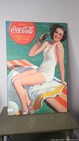 Photo of mounted vintage Coca-Cola advertisement poster showing a woman in a white swimsuit holding a Coke bottle, with the iconic Coca-Cola logo and slogan in red circle design.