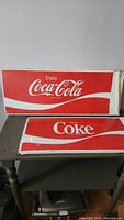 Front view of two rectangular Coca-Cola metal signs on a table, showing brand text and red-white wave design