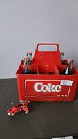 Red plastic Coca-Cola bottle holder containing four glass bottles of Coca-Cola with metal caps and Coca-Cola branding.