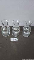Three clear glass Coca-Cola shot glasses, each with a flared top and white Coca-Cola logo printed on the front, displayed on a dark surface.