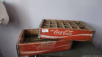 Two wooden Coca-Cola trays stacked, showing red painted sides with Coca-Cola logo and metal-reinforced corners.
