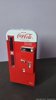 Front view of the red and white Coca-Cola toy bottle dispenser resembling a vintage vending machine with coin slot and bottle dispensing feature.