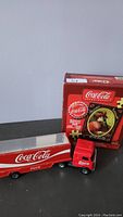 Photo showing Coca-Cola puzzle box standing upright behind the red and white Coca-Cola toy truck with trailer on a dark surface.