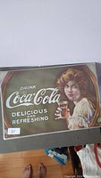 Metal place mat with vintage Coca-Cola logo and woman holding glass of Coca-Cola, text 'Drink Coca-Cola Delicious and Refreshing'