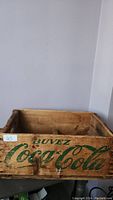 Front side view of the rectangular wooden crate showing the Coca-Cola logo in green paint and the word 'Buvez' above it. The crate is visibly aged with wear marks.
