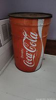 Full side view of orange/red cylindrical metal bin with white Coca-Cola logo lettering on side, showing wear and scratches.