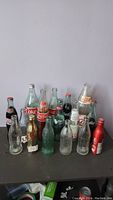 Photo showing 16 Coca-Cola bottles arranged in staggered rows, various glass and aluminum bottles with vintage and classic labels.