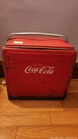 Front view of red metal cooler with Coca-Cola logo and white handle on top, showing signs of wear and scratches.