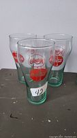 Three Coca-Cola glasses showing red bottle cap logos, all clear glass with slight green tint, displayed on metal surface.